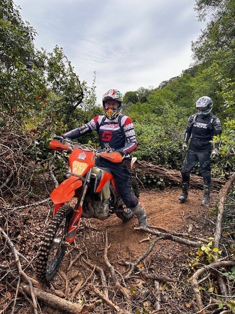 Two enduro riders on a wooded trail with exposed roots and fallen branches